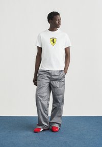 Λευκό T-shirt με κίτρινο λογότυπο της Ferrari, συνδυασμένο με φαρδιά, γυαλιστερή γκρι παντελόνα και κόκκινα παπούτσια Puma slip-on, που στέκεται σε μπλε χαλί.