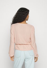 Camisola longa de manga rosa texturada com cintura franzida, apresentando um padrão subtil. Combinada com jeans azul claro.