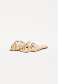 Gylne sandaler med flate såler har et fletet stroppdesign og firkantet tå. Laget av et blankt materiale med en glatt tekstur.