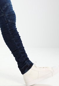 Mörkblå, rynkade skinny jeans i kombination med vita högtoppsskor, mot en ljus bakgrund.