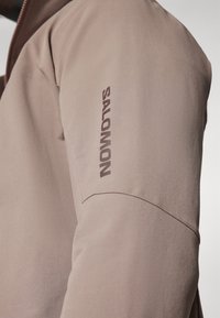 Svetlo rjava flis jakna z teksturirano tkanino, ki ima tesno prilegan dizajn in temno rjav "SALOMON" logotip na rokavu.