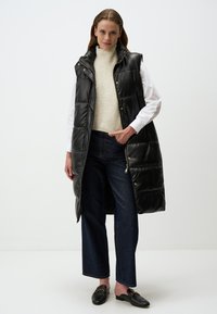 Gilet sans manches rembourré noir avec une texture brillante, col montant et boutons-pression, porté sur un pull en tricot crème et un jean évasé bleu.