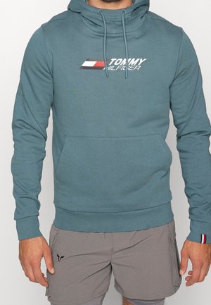 Mann, der einen türkisen Tommy Hilfiger Hoodie und graue Shorts trägt, mit entspannt an den Seiten hängenden Händen, vor einem einfarbigen Hintergrund stehend.