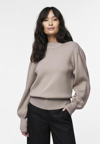 Pull léger beige avec un col rond, des manches bouffantes, et des bords ribblés. Porté avec un pantalon large noir.