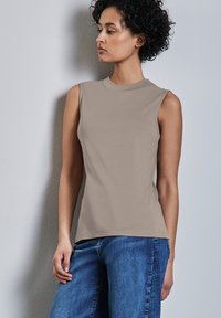 Donna con capelli ricci che indossa un top senza maniche color taupe e jeans blu, in piedi contro un muro grigio chiaro, mentre guarda di lato.