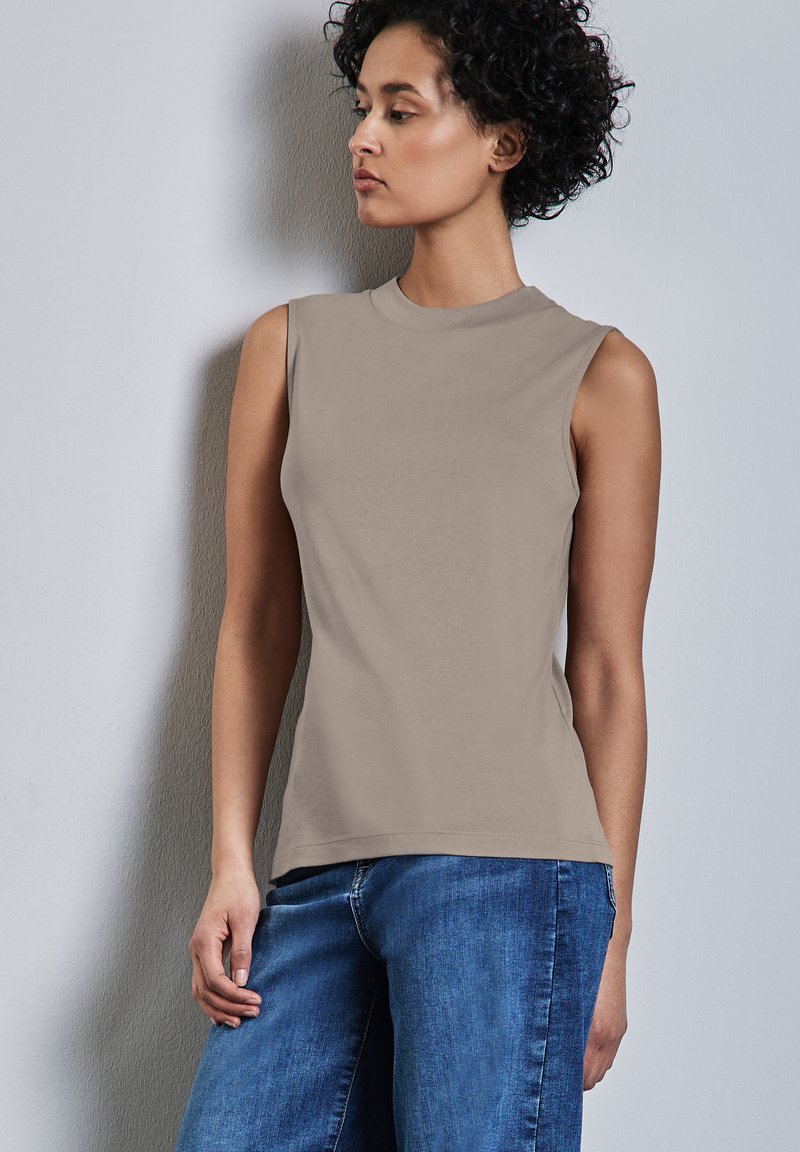 Donna con capelli ricci che indossa un top senza maniche color taupe e jeans blu, in piedi contro un muro grigio chiaro, mentre guarda di lato.
