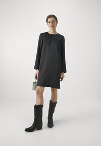 Abito lungo a maniche nere con dettagli a pannello curvo, abbinato a stivali da cowboy neri alti. La modella tiene una piccola borsa scintillante verde.