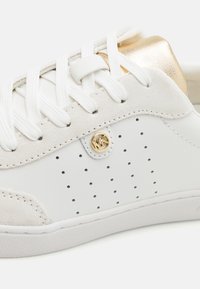 Baskets blanches avec un bout en daim texturé, design perforé, lacets blancs plats et un accent doré métallique à l'arrière. Détails du logo doré visibles.