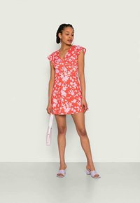 Vestido floral vermelho com mangas curtas e volantes, decote em V e forma ajustada. Usado com acessórios em lavanda, incluindo uma bolsa e sapatos.