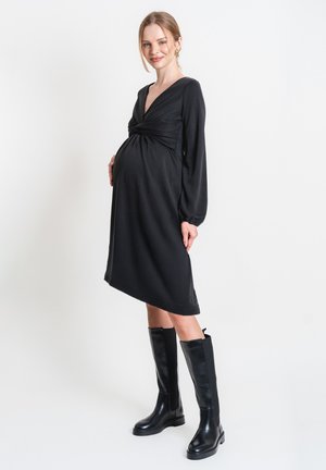 Zwangere vrouw staand, gekleed in een zwart, lang shirt met lange mouwen en zwarte kniehoge laarzen, glimlachend met één hand op haar buik.