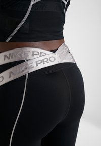 Czarne legginsy sportowe z białymi detalami ściegów, z szerokim, odblaskowym pasem oznaczonym "NIKE PRO". Gładki, elastyczny materiał.
