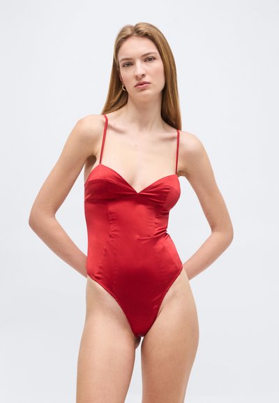 Štíhlá žena s rovnými blond vlasy, oblečená v červených saténových jednodílných plavkách s tenkými ramínky, pózující před jednoduchým světlým pozadím.