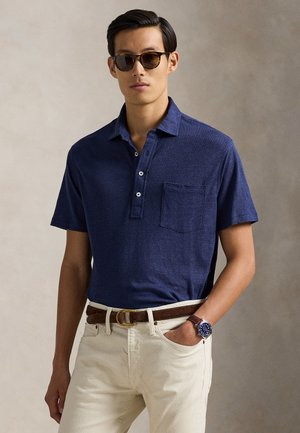 Polo blu navy con patta a tre bottoni e taschino; abbinata a pantaloni chiari e cintura marrone; indossa occhiali da sole.