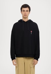 Felpa nera con vestibilità comoda, realizzata in tessuto morbido. Presenta un piccolo design ricamato a forma di lecca-lecca sul petto, con cordini nel cappuccio.