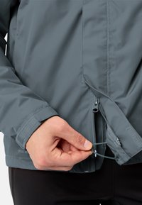 Giacca grigia impermeabile con finitura strutturata. Presenta molteplici cerniere, cordino regolabile all'orlo e polsini su misura per una vestibilità sicura.