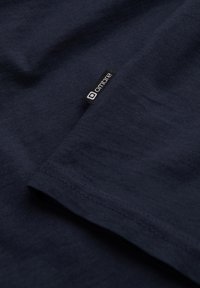 Marineblaue Baumwoll-T-Shirt mit glatter Textur, versehen mit einem schwarzen Etikett mit weißem Text "ombre" nahe dem Saum. Flach anliegendes Ärmeldetail sichtbar.