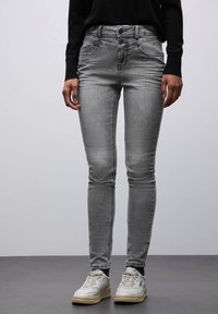 Jeans skinny de mezclilla gris con cintura alta, que presentan un cierre de dos botones y bolsillos frontales. Combinados con zapatillas blancas.