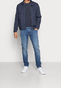 Marinblå bomberjacka med två framfickor, bärs över en texturerad marinblå stickad tröja, ihop med ljusblå jeans och vita sneakers.