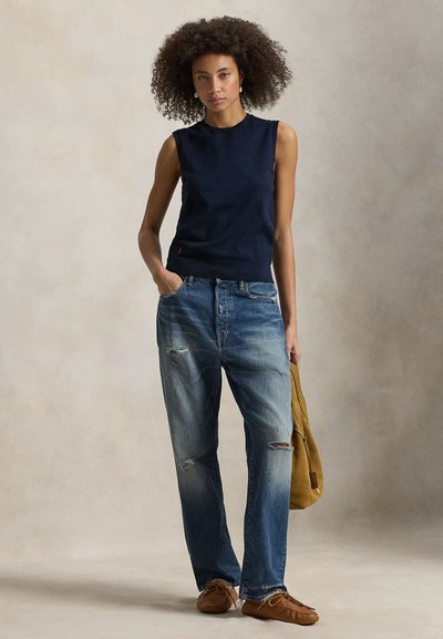 Femme aux cheveux bouclés portant un top sans manches bleu marine, un jean ample déchiré, des mocassins marron et tenant un sac en daim beige.