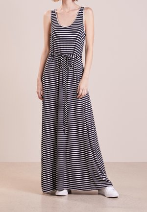 Femme portant une robe longue sans manches à rayures horizontales noires et blanches avec une taille cintrée et des baskets blanches, debout sur un sol beige.