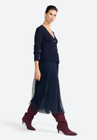 Marineblauwe geribde cardigan met zes knopen, gelaagd over een geplisseerde, doorschijnende marineblauwe rok. Bordeaurode suède kniehoge laarzen met spitse neuzen maken de outfit compleet.