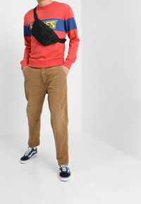 Červená mikina s modrými pruhovanými akcenty a logem, béžové roztrhané kalhoty, černá crossbody taška a modré tenisky s bílou podešví.