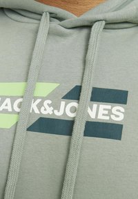 Ljusgrå huvtröja i ett texturerat tyg, med en vit logotyp och gröna samt mörkblå geometriska accenter. Dragsnörena är i matchande färg.