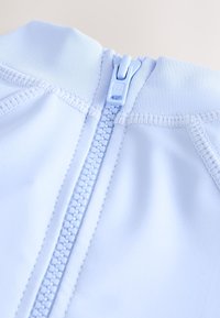 Lichtblauwe stof met een gladde textuur, voorzien van een centraal geplaatste rits en gestikte details langs de randen. Het ritssluitinkje is ook lichtblauw.