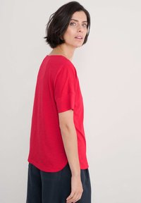 Rotes T-Shirt mit kurzen Ärmeln und rundem Halsausschnitt, in entspannter Passform. Es hat eine glatte Textur und einen abgerundeten Saum. Kombiniert mit dunklen Hosen.