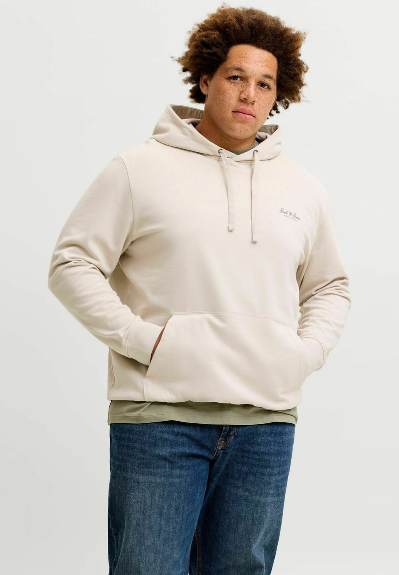 Jeune homme aux cheveux bouclés portant un sweat à capuche beige et un jean bleu, debout les mains dans la poche du sweat sur un fond uni.