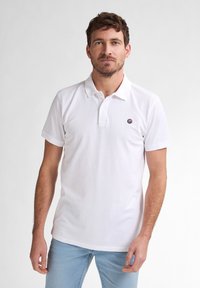 Polo bianco in cotone con colletto a bottoni, maniche corte e un piccolo logo ricamato sul petto. Indossato con jeans azzurri chiari.