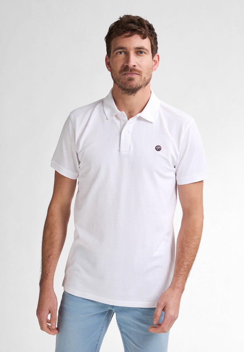 Polo bianco in cotone con colletto a bottoni, maniche corte e un piccolo logo ricamato sul petto. Indossato con jeans azzurri chiari.
