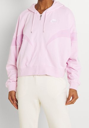 Osoba ubrana w jasnoróżową bluzę Nike na zamek z kapturem i białym logo Nike Air, w połączeniu z wysokim stanem kremowych spodni.