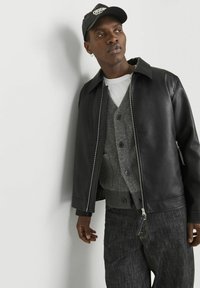 Black leather jacket with a front zipper, worn over a gray buttoned cardigan and white t-shirt; paired with dark textured pants and a cap.