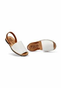 Sandalias de cuero blanco tipo slip-on con una correa marrón y suela de goma negra. Presentan una textura suave y un diseño de punta redondeada.
