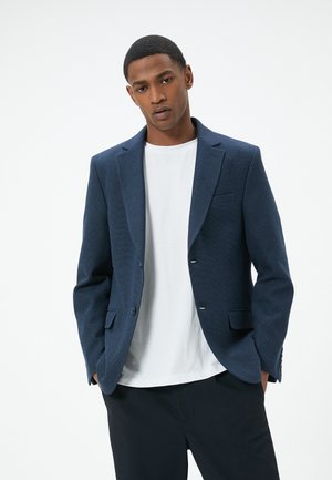 Marineblauer strukturierten Blazer mit slim fit, einreihigem Design, Reverskragen und zwei vorderen Taschen, getragen über einem weißen Rundhals-Shirt.
