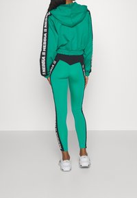 Felpa verde con accenti neri e bordi bianchi del logo, abbinata a leggings verdi con strisce laterali nere e una vita a contrasto. Sneakers bianche.