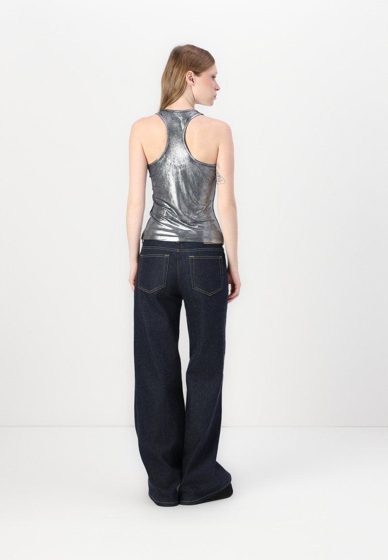 Silberfarbener, metallischer ärmelloser Oberteil, kombiniert mit dunkelblauen, weit geschnittenen Jeans. Der Oberteil hat ein tailliertes Design, während die Jeans über traditionelle Gesäßtaschen verfügen.
