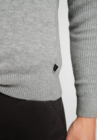 Šedý pletený svetřík s žebrovanými manžetami a lemem. Obsahuje malou černou nášivku na boku. Vyroben z měkké, texturované látky.