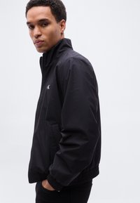 Μαύρο μπουφάν τύπου bomber με φερμουάρ, ελαφρύ ύφασμα, ριμπ μανσέτες και τελείωμα, διακριτική τσέπη, λογότυπο στο στήθος, λεία υφή, casual σχεδιασμός.