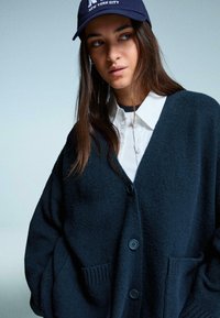 Tmavě modrý pletený kardigan s výstřihem do V, vpředu na knoflíky a dvěma předními kapsami, nošený přes bílou košili s límečkem a námořnickou čepicí.