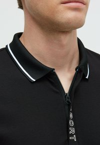 Polo negro con cremallera en la parte delantera, que presenta un cuello texturizado y rayas blancas. La tela es suave, con un detalle impreso de "SPORT".