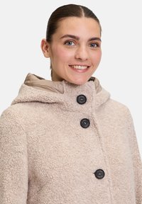 Beige Fleece-Mantel mit strukturiertem Obermaterial, ausgestattet mit einer Kapuze, einem kontrastierenden hellen Innenfutter und drei schwarzen Knöpfen auf der Vorderseite.
