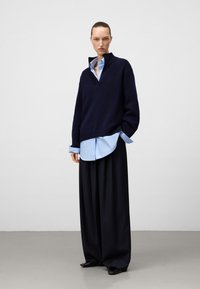 Maglione blu navy oversize con colletto alto indossato sopra una camicia azzurro chiaro con bottoni e pantaloni neri larghi, esposto su uno sfondo neutro.