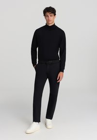 Maglione a collo alto nero, pantaloni neri slim-fit, cintura nera texturizzata e sneakers bianche. Tessuto morbido, taglio moderno e stile minimale.