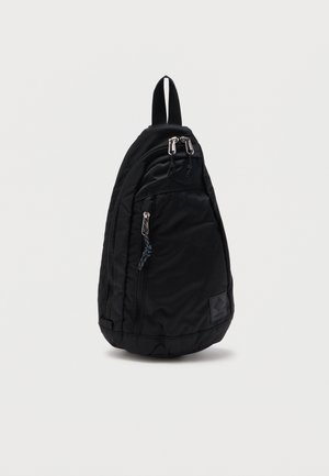 Sac à dos bandoulière noir à une sangle avec poches avant zippées et une boucle de transport supérieure sur fond blanc.