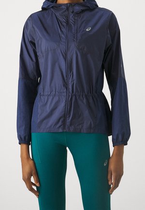 Persoon draagt een marineblauwe capuchonjas met elastische manchetten en teal leggings, staande tegen een effen lichte achtergrond.