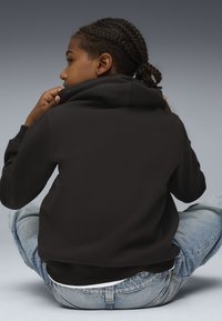 Felpa nera con cappuccio dal taglio rilassato, orlo e polsini a costine. Persona seduta a gambe incrociate, indossando jeans azzurri strappati.