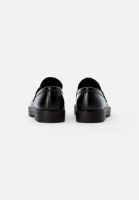 Chaussures en cuir noir avec une surface lisse, des talons arrondis et une semelle en caoutchouc basse. Elles présentent des coutures minimales et aucun matériel visible.