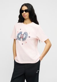 Γυναίκα με μακριά σκούρα μαλλιά που φοράει μαύρα γυαλιά ηλίου, ανοιχτό ροζ μπλουζάκι με σχέδιο ήλιου και σελήνης, και μαύρο παντελόνι με τα χέρια στις τσέπες.